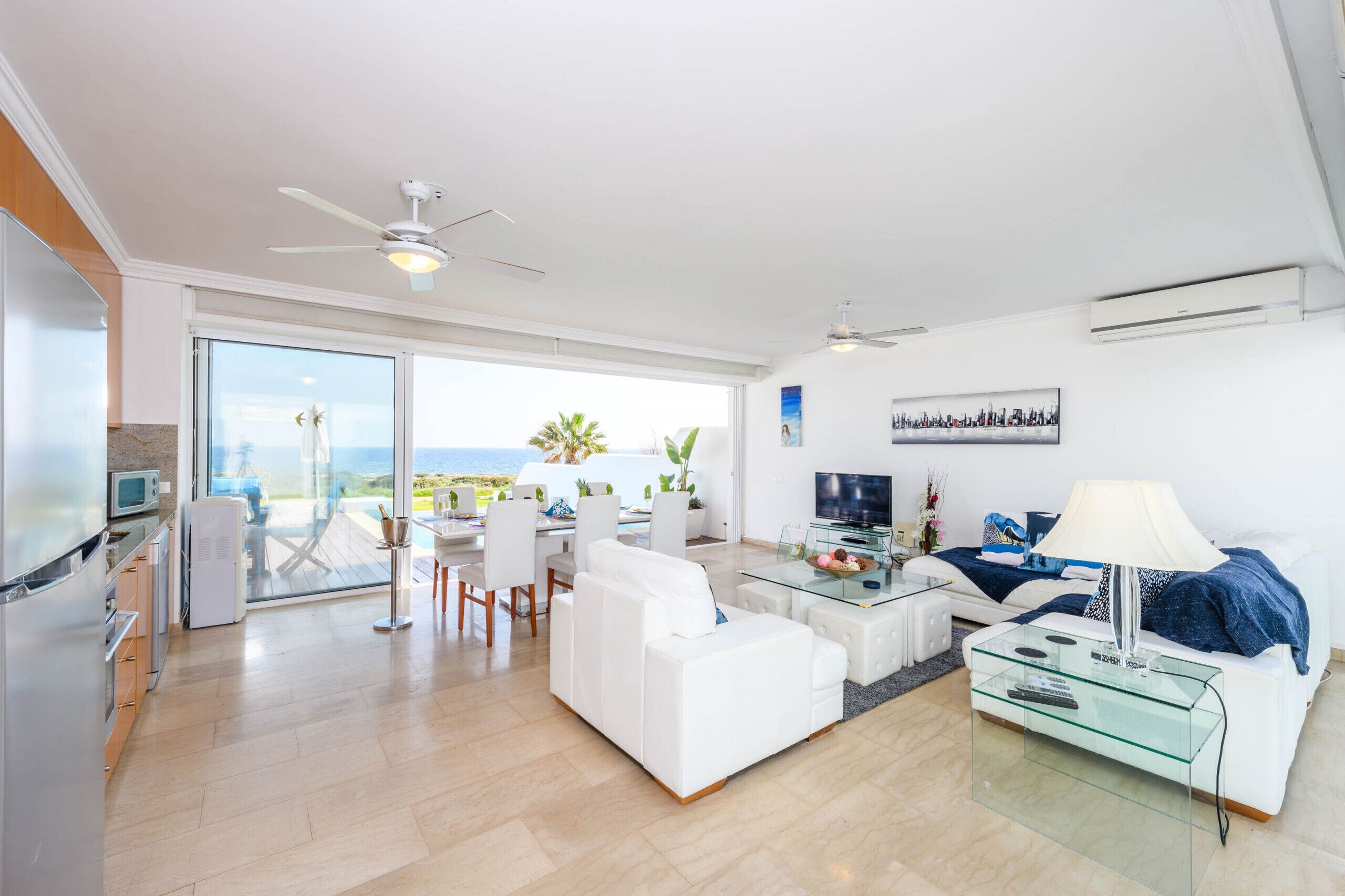 A modern living room with a white sofa, a glass coffee table, and a TV. There is a balcony with a view of the ocean.