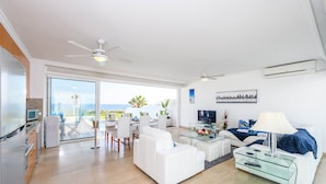 Una sala de estar moderna con un sofá blanco, una mesa de centro de vidrio y una televisión. Hay un balcón con vista al océano.