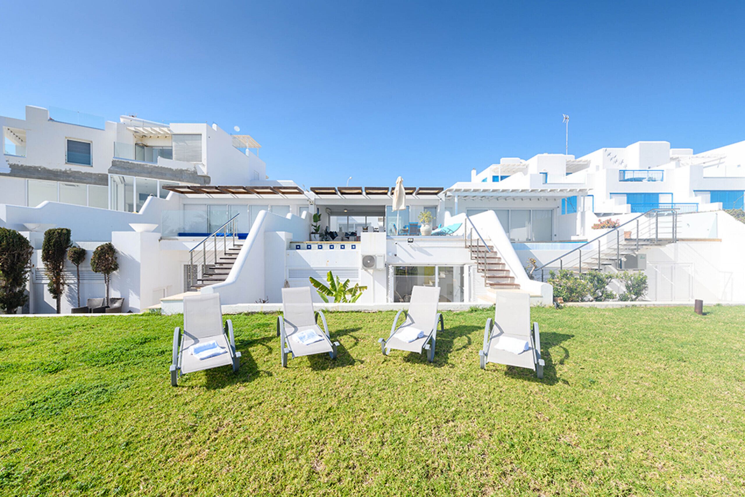 Un bâtiment moderne de couleur blanche, doté d’un vaste espace extérieur comprenant quatre chaises longues et un escalier menant à un niveau supérieur.