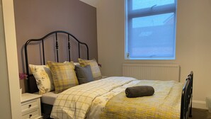A neatly made bed with a patterned bedspread and pillows, a bedside table with a lamp, and a window with a view of the outside. - Gordon House. (Kettering)