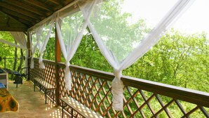 A covered outdoor area with a wooden deck, white curtains, and a view of green trees.