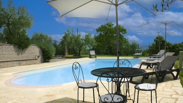A swimming pool with lounge chairs and a table, surrounded by a stone wall and trees.