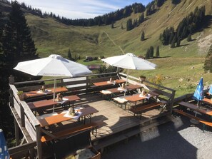 Eine Außenterrasse mit Holztischen und -bänken, weißen Sonnenschirmen und Blick auf eine Berglandschaft.