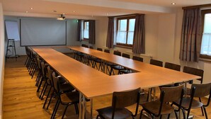 Ein Konferenzraum mit langem Tisch und Stühlen, einer Projektionsleinwand und Fenstern mit Vorhängen.