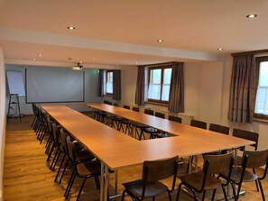 Ein Konferenzraum mit langem Tisch und Stühlen, einer Projektionsleinwand und Fenstern mit Vorhängen.