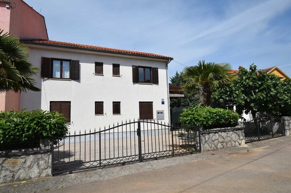 A two-story building with a white facade and brown shutters, a stone wall, and a metal gate.