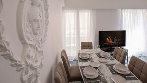 Un comedor con una mesa de mármol dispuesta para seis personas, rodeada de sillas tapizadas. Un televisor está montado en la pared y la habitación tiene paredes blancas y ventanas grandes con cortinas de gasa.