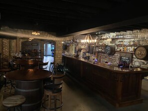 Un espace bar avec des tables et des tabourets en bois, un mur en pierre et une variété de bouteilles et de verres exposés sur des étagères.