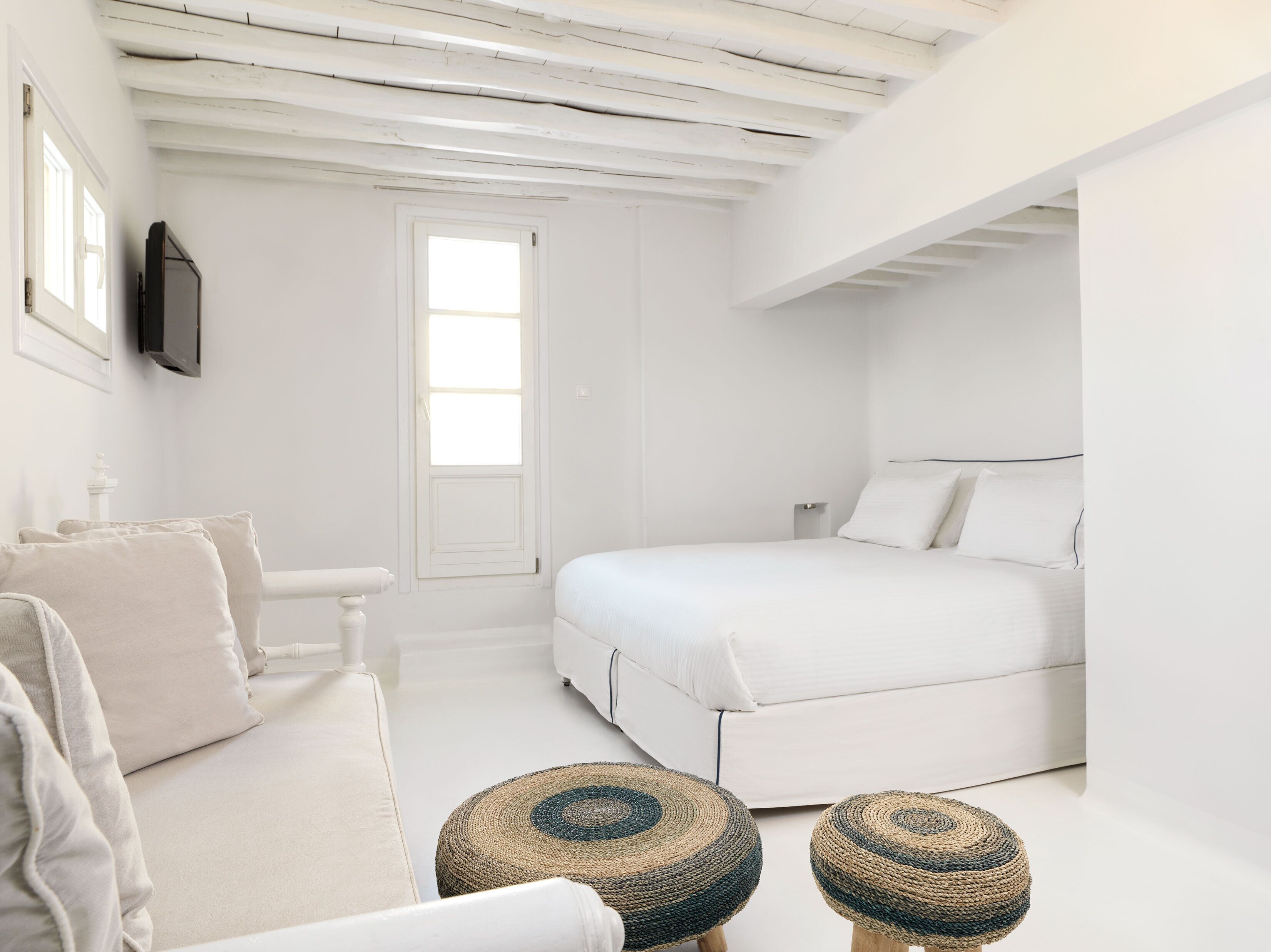 A minimalist white bedroom with a bed, a small white table, and two woven stools.