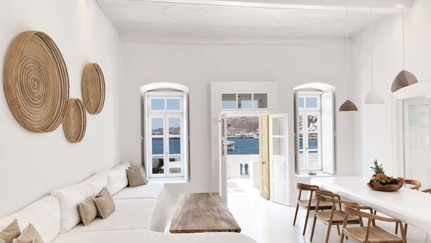 Ett rymligt vardagsrum med en vit soffa, ett matbord med trästolar och utsikt över havet genom stora fönster.
