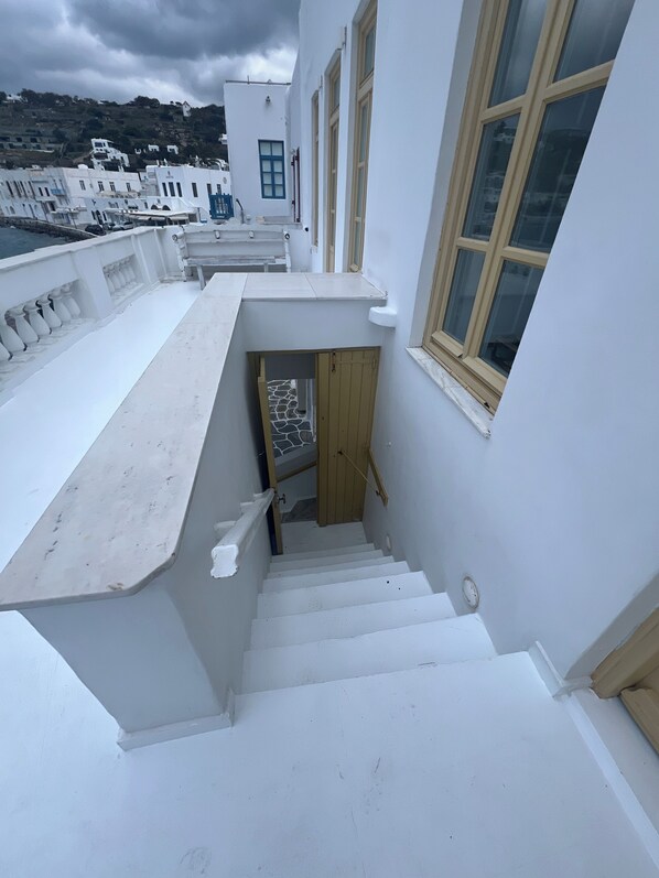 Una escalera blanca que conduce a una puerta, con una pared de piedra de fondo, rodeada de edificios blancos con ventanas amplias.