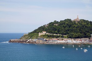 Un pueblo costero con una estatua destacada en la cima de una colina, una estructura fortificada y un puerto deportivo con barcos.