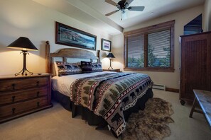 A bedroom with a bed, two bedside lamps, a ceiling fan, a window with blinds, and a wooden dresser.