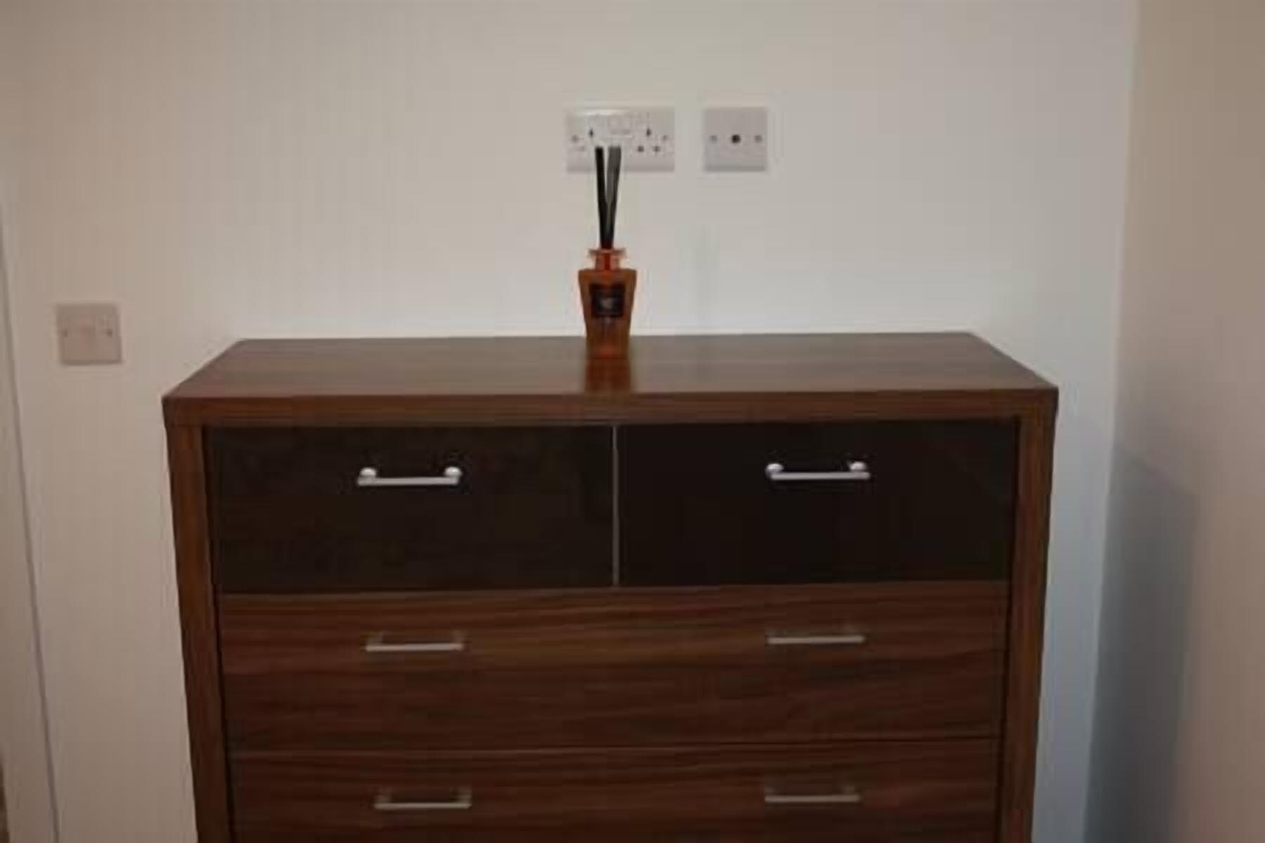 A wooden cabinet with two drawers and a shelf, placed against a plain wall with two electrical outlets.