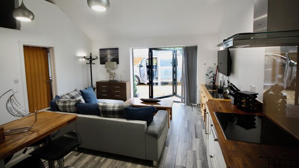 A modern kitchen with a wooden countertop, a built-in oven, and a stainless steel range hood. There is a dining area with a wooden table and chairs, and a sofa with plaid cushions.