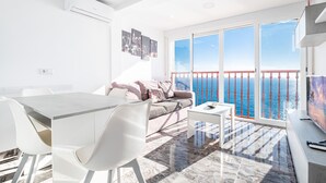 Una sala de estar moderna con un balcón que da al mar, un comedor con muebles blancos y un televisor grande a la derecha.