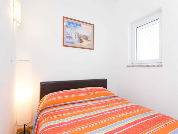 A single bed with a striped bedspread, a bedside lamp, a framed painting on the wall, and a window with blinds. - Apartment Josip 2 with Seaview (Čižići)