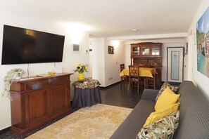 A living room with a flat-screen TV, a wooden cabinet, a sofa with yellow cushions, a dining area with a table and chairs, and a kitchen area with a cabinet.