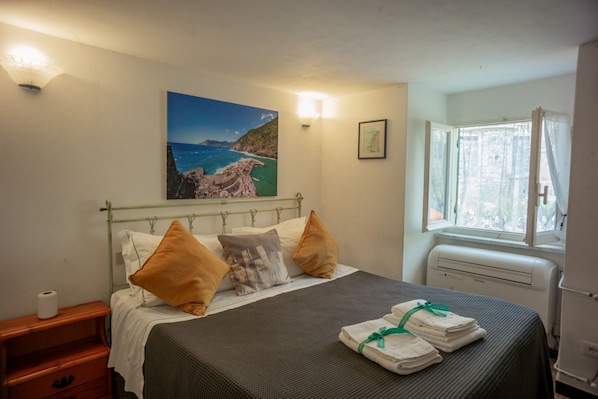 A neatly made bed with a view of the sea, a bedside table with a lamp, and a window with curtains.