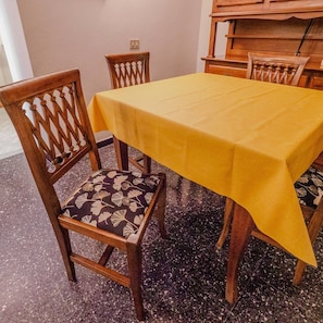 Un comedor con mantel amarillo, sillas de madera con cojines estampados y piso de baldosas.