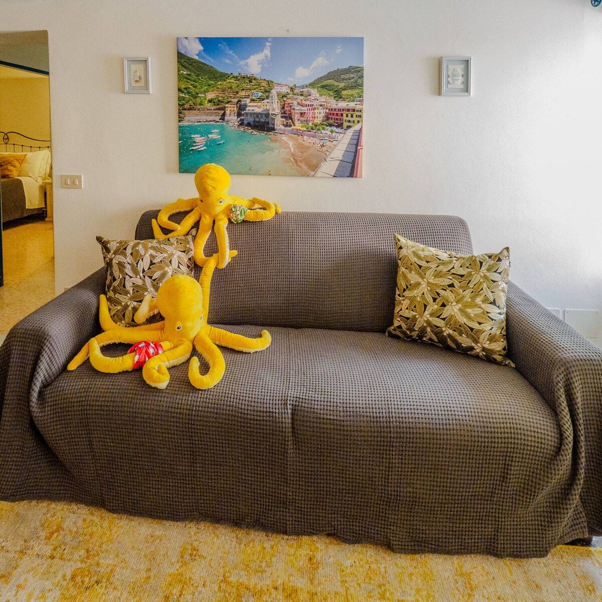 A grey sofa with a large octopus plush toy and patterned pillows, a framed picture of a coastal town, and a yellow carpet.