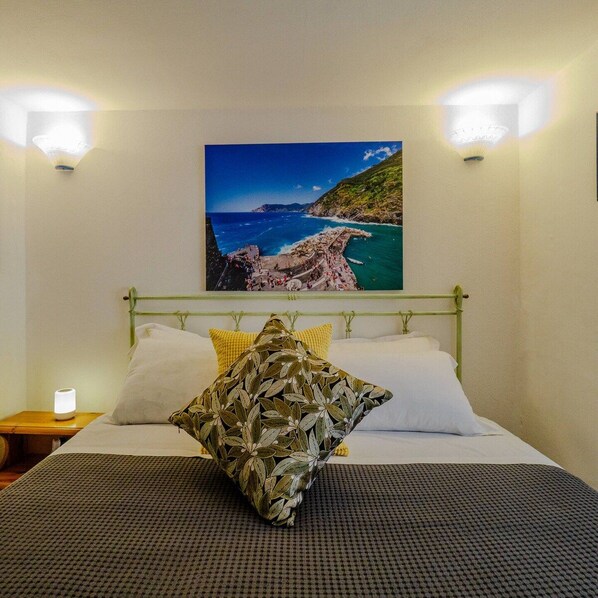 A bed with a patterned pillow, a nightstand with a lamp, and a wall-mounted picture of a coastal landscape.