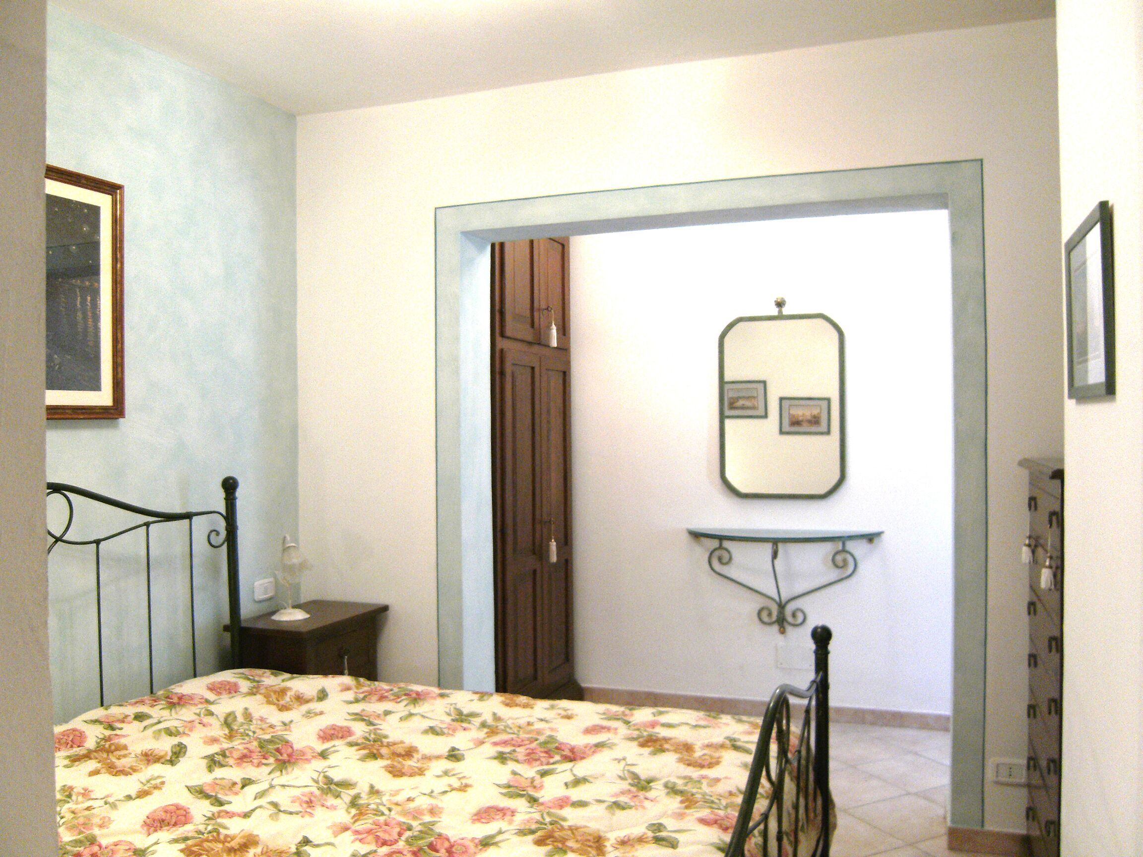 Ein Schlafzimmer mit blumigem Bettbezug, einem Nachttisch, einem Spiegel an der Wand und einer Tür zu einem anderen Raum.