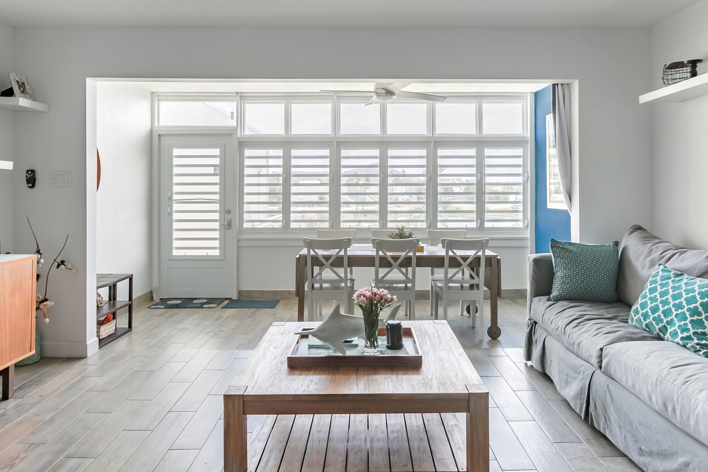 A bright living room with a wooden coffee table, a grey sofa, and a dining area with a table and chairs.