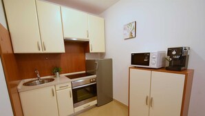 Una cocina compacta con armarios blancos, un lavamanos de acero inoxidable, un horno, un refrigerador, un microondas y una cafetera.