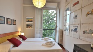 Ein Hotelzimmer mit zwei Betten, einem Balkon mit Blick auf Grünflächen und einer Wand, die mit Vintage-Automobilplakaten dekoriert ist.