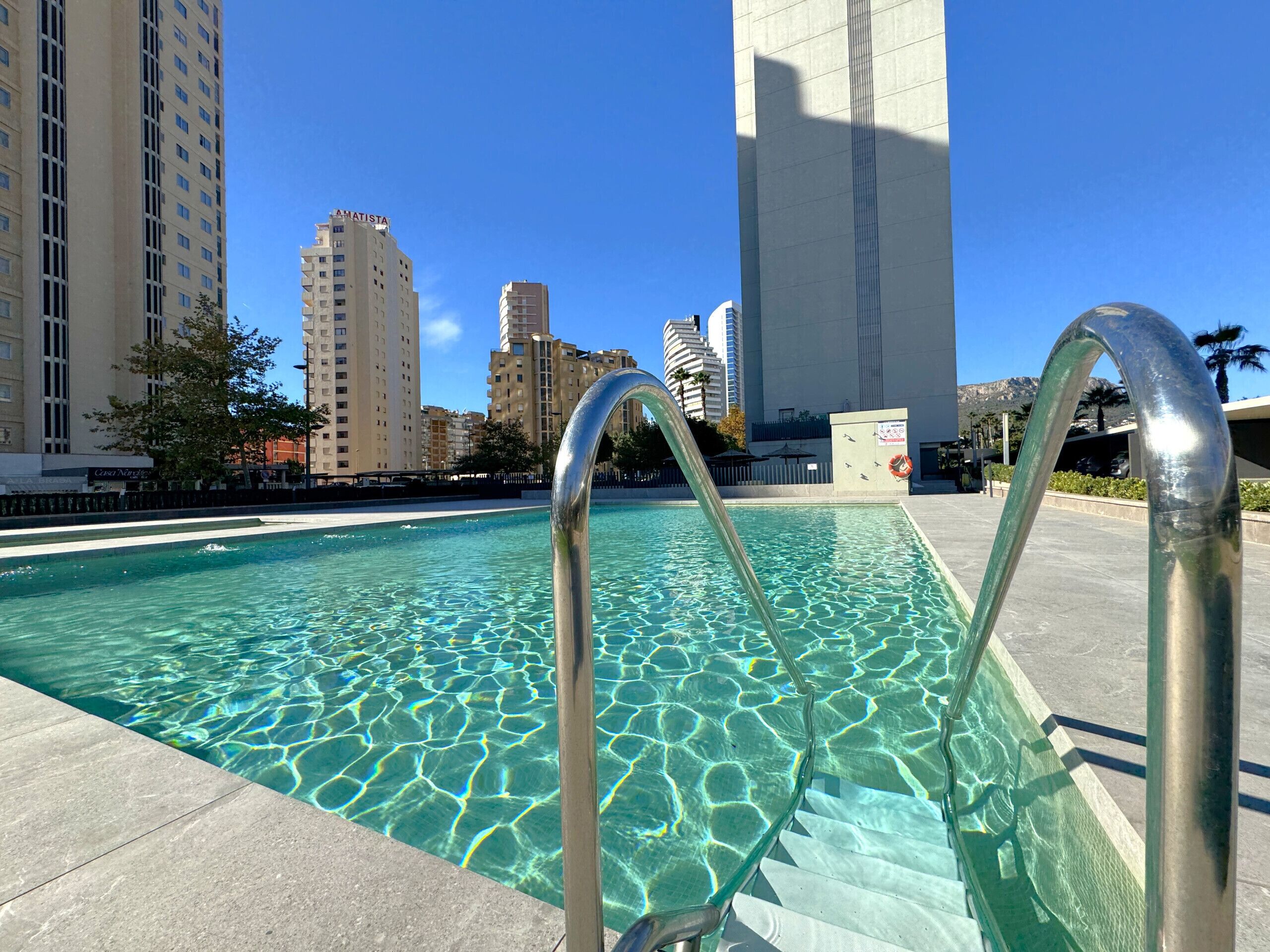A swimming pool with clear water and metal pool ladders, surrounded by tall buildings and a clear sky.
