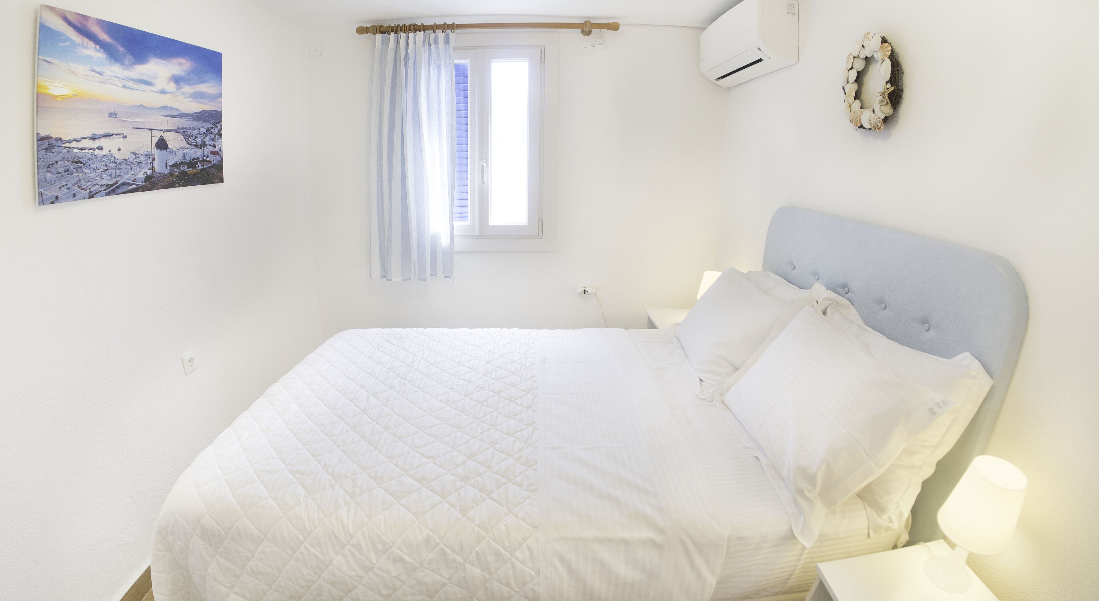 Um quarto com cama, janela com cortinas, ar-condicionado e um objeto decorativo na parede.