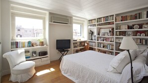 Quarto com cama, uma cadeira, televisão e estante cheia de livros e objetos decorativos.