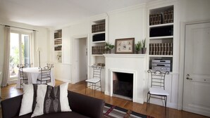 Uma sala de estar com lareira, estantes de livros, uma área de jantar com mesa e cadeiras, e um sofá com almofadas estampadas.