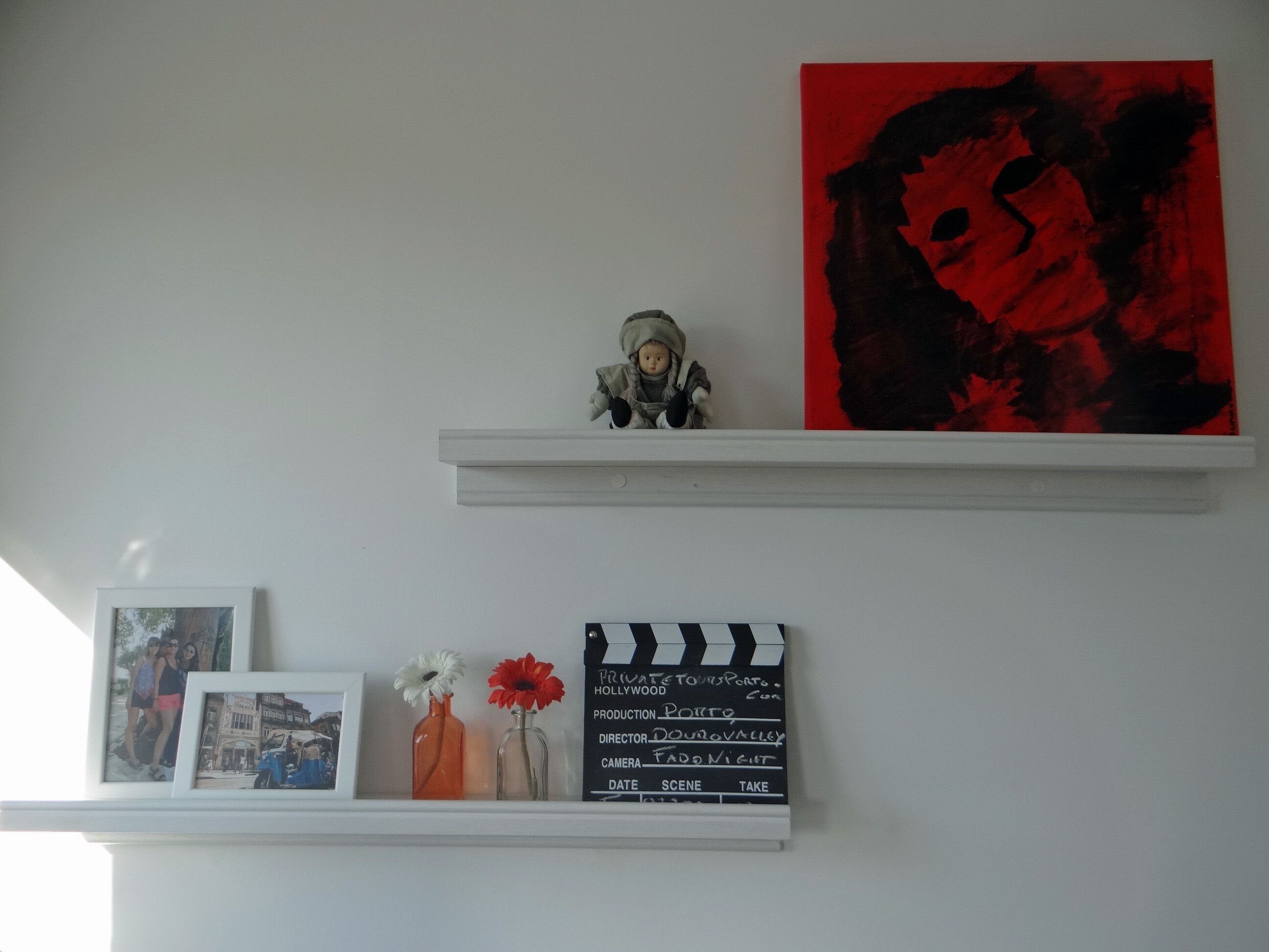 Eine Wand mit einem weißen Regal, das ein Filmklappe, zwei Vasen mit Blumen und ein gerahmtes Foto zeigt.