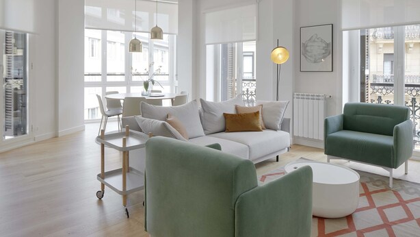 A modern living room with a white sofa, green armchairs, and a coffee table. There is a dining area with a table and chairs, and a view of the city through large windows. - Belle Sebastian by FeelFree Rentals (San Sebastián)
