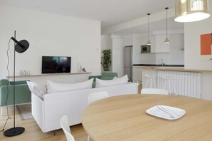 A modern living room with a sofa, a TV, a dining table, and a kitchen area in the background.