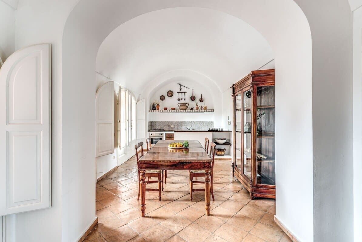 Une cuisine spacieuse avec une grande table en bois, des meubles blancs et un sol carrelé.