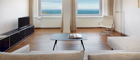 A modern hotel room with a large window offering a view of the sea, a flat-screen TV, a small table, and two sofas.
