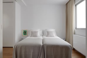A modern hotel room with two beds, a large window with curtains, and a minimalist design.