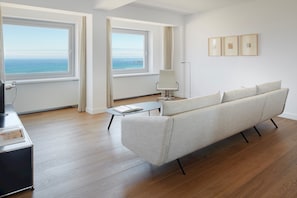 A modern living room with a large sofa, a coffee table, and a chair. There are two windows with ocean views.