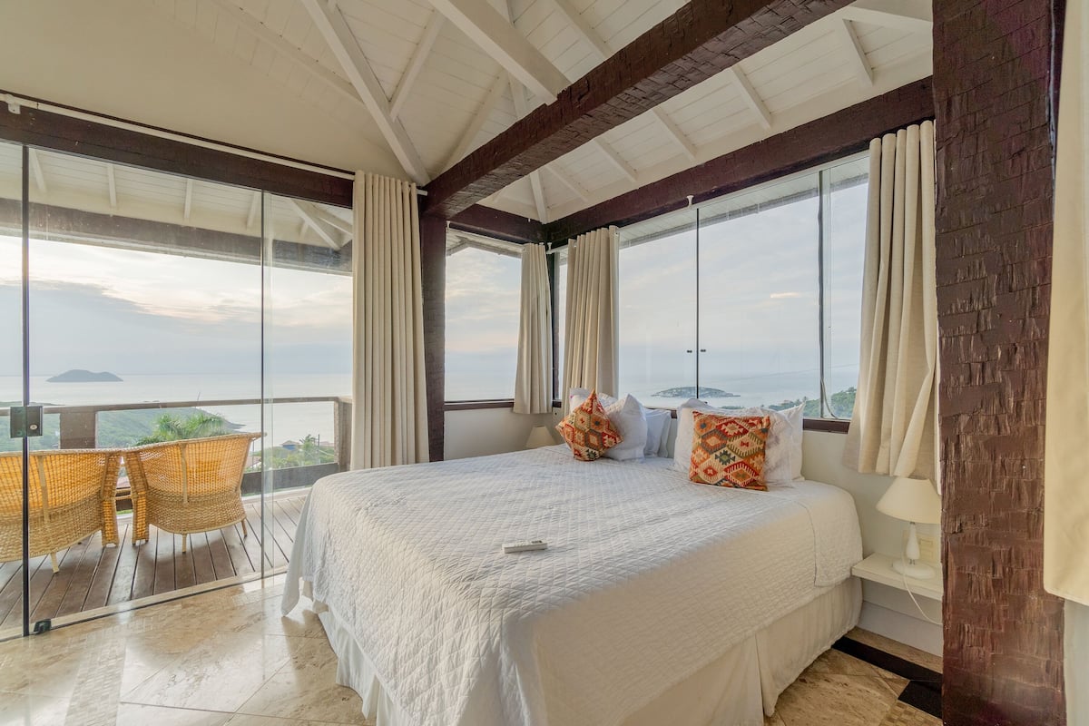 Ein Schlafzimmer mit einem großen Bett, einem Balkon mit Korbstühlen und Blick auf das Meer.