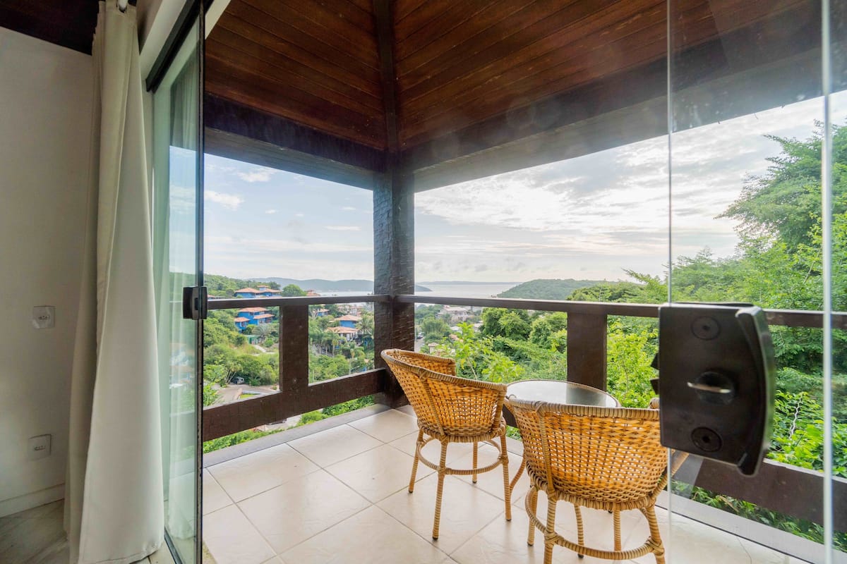 Ein Balkon mit Korbstühlen und einem Tisch, mit Blick auf eine üppige grüne Landschaft und entfernte Gebäude.