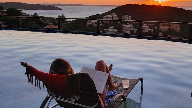 A person relaxing on a lounge chair with a book, overlooking a sunset view with hills and a body of water. - Eny Boutique Hotel (Búzios)