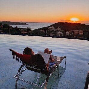 A person relaxing on a lounge chair with a book, overlooking a sunset view with hills and a body of water. - Eny Boutique Hotel (Búzios)