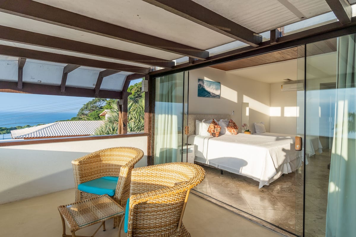 Ein Balkon mit Korbmöbeln, ein Bett mit weißer Bettwäsche und eine Glaswand mit Blick aufs Meer.