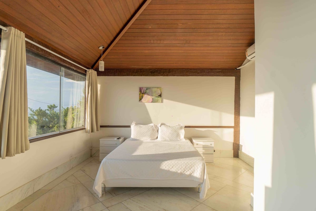 Ein Schlafzimmer mit einem großen Bett, Holzboden, einem Fenster mit Vorhängen, einem Nachttisch und einem Beistelltisch.