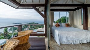 A balcony with wicker furniture, a bed with white bedding, and a view of the sea and greenery. - Eny Boutique Hotel (Búzios)