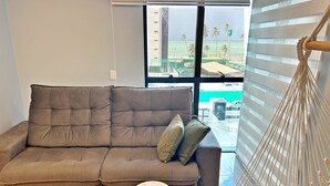 Een moderne woonkamer met een grijze bank, twee kleine houten krukjes, een macramé hangstoel en uitzicht op een strand en palmbomen door grote ramen.