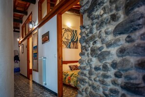 Un couloir avec un mur en pierre, des poutres en bois, un radiateur et un lit recouvert d’une couvre-lit à motifs floraux.